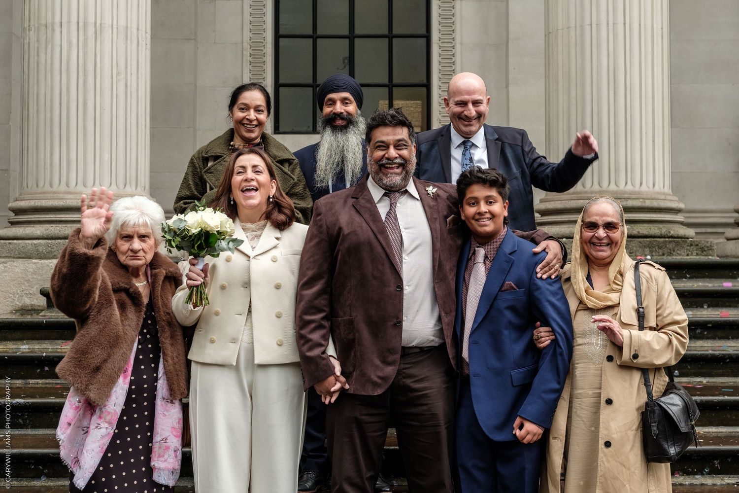 A joyful group of eight family and friends celebrate on courthouse steps, smiling and waving, with a woman holding a white bouquet.