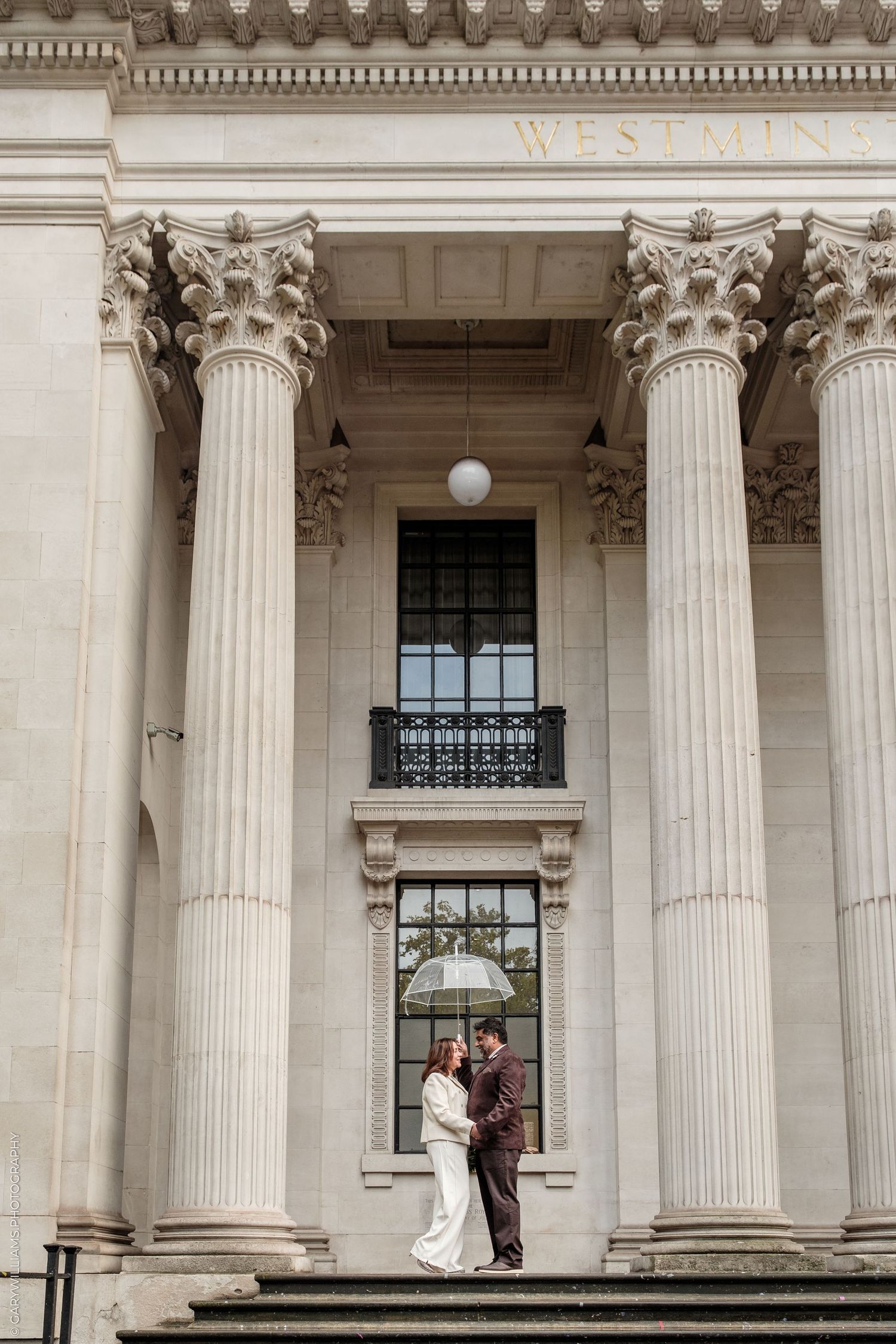 Couple embracing under a clear umbrella on the steps of a grand neoclassical building with tall Corinthian columns.
