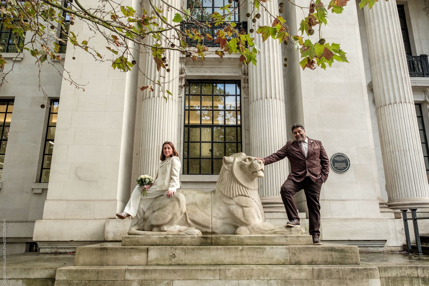 Bride in a white dress sits on a stone lion statue while the groom in a brown suit stands beside her outside a neoclassical building.