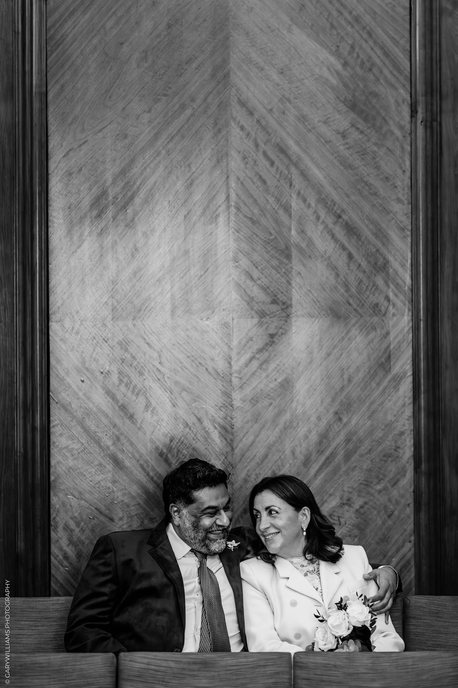 Black and white photo of a smiling couple seated close together, the bride holding a white bouquet, against a herringbone wood-paneled wall.