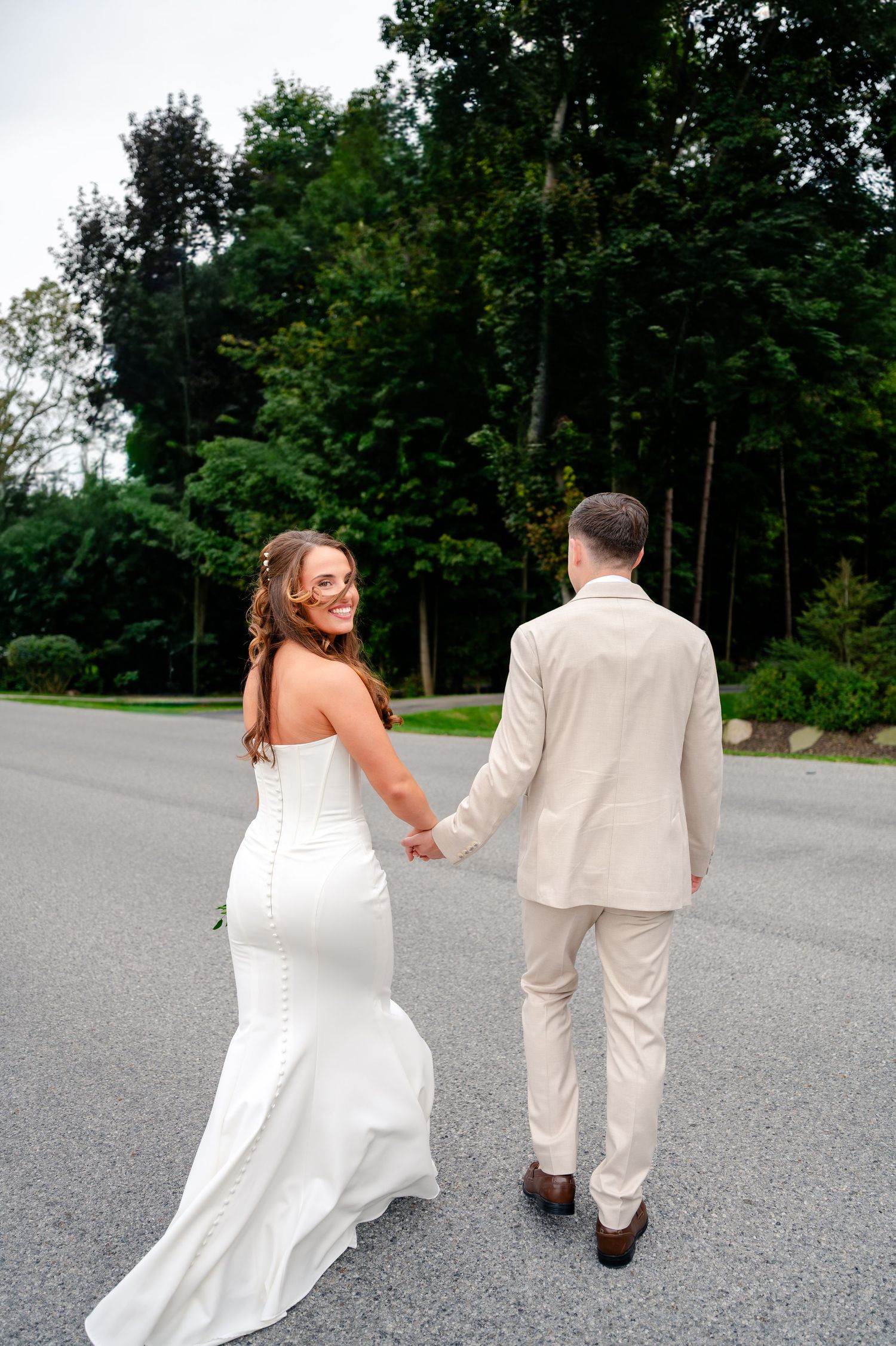 Bride in a white fitted gown smiles over her shoulder while holding hands with the groom in a cream suit on a tree-lined road.