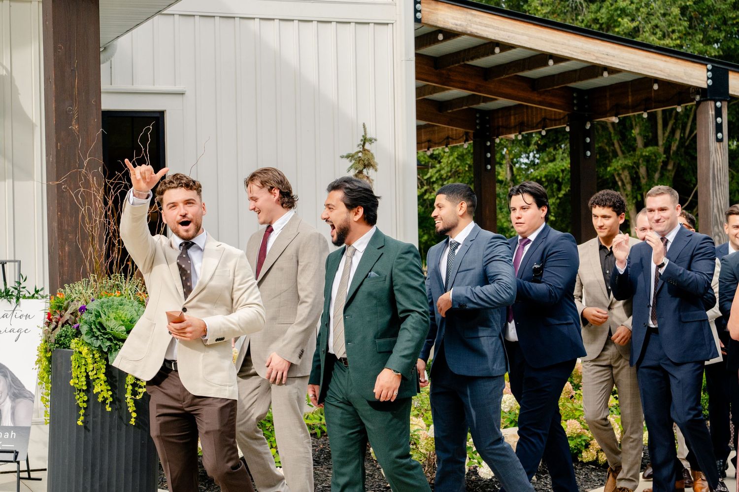 Groomsmen in green, tan, and navy suits walk together outdoors at a wedding, smiling and cheering beside a white building with greenery.