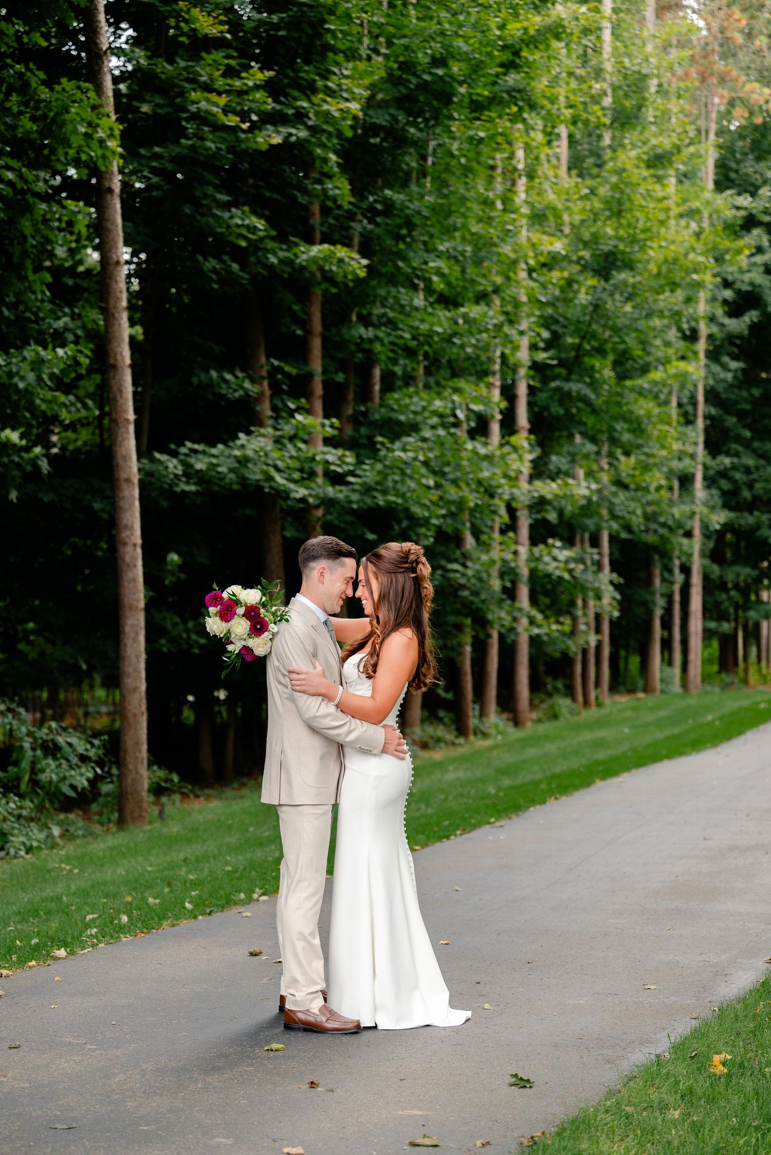 Bride and groom embrace on a paved path lined with tall green trees, she in a fitted white gown holding a bouquet of pink and white flowers.