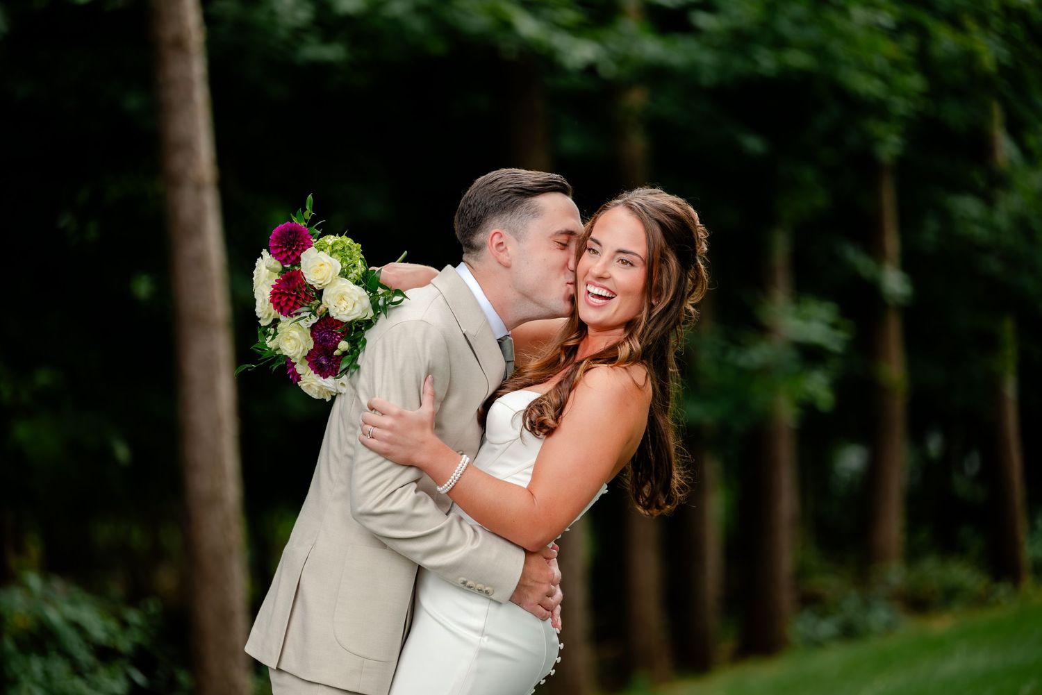 Groom in a tan suit kisses a smiling bride in a white dress holding a bouquet of burgundy and white flowers outdoors among green trees.