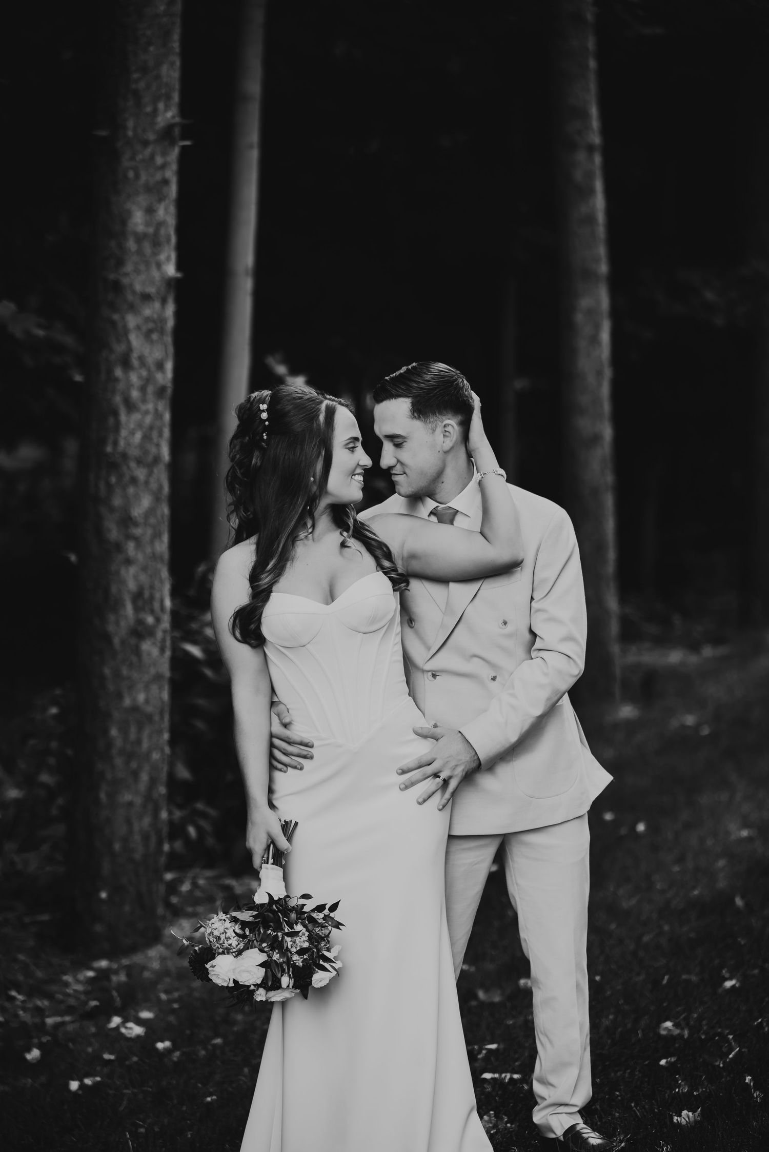 Black and white photo of a bride in a fitted gown and groom in a light suit embracing among tall trees in a forest.