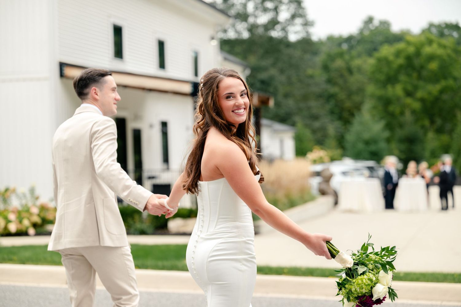 Smiling bride in a white dress holds a green bouquet and her groom's hand while walking outdoors toward wedding guests.