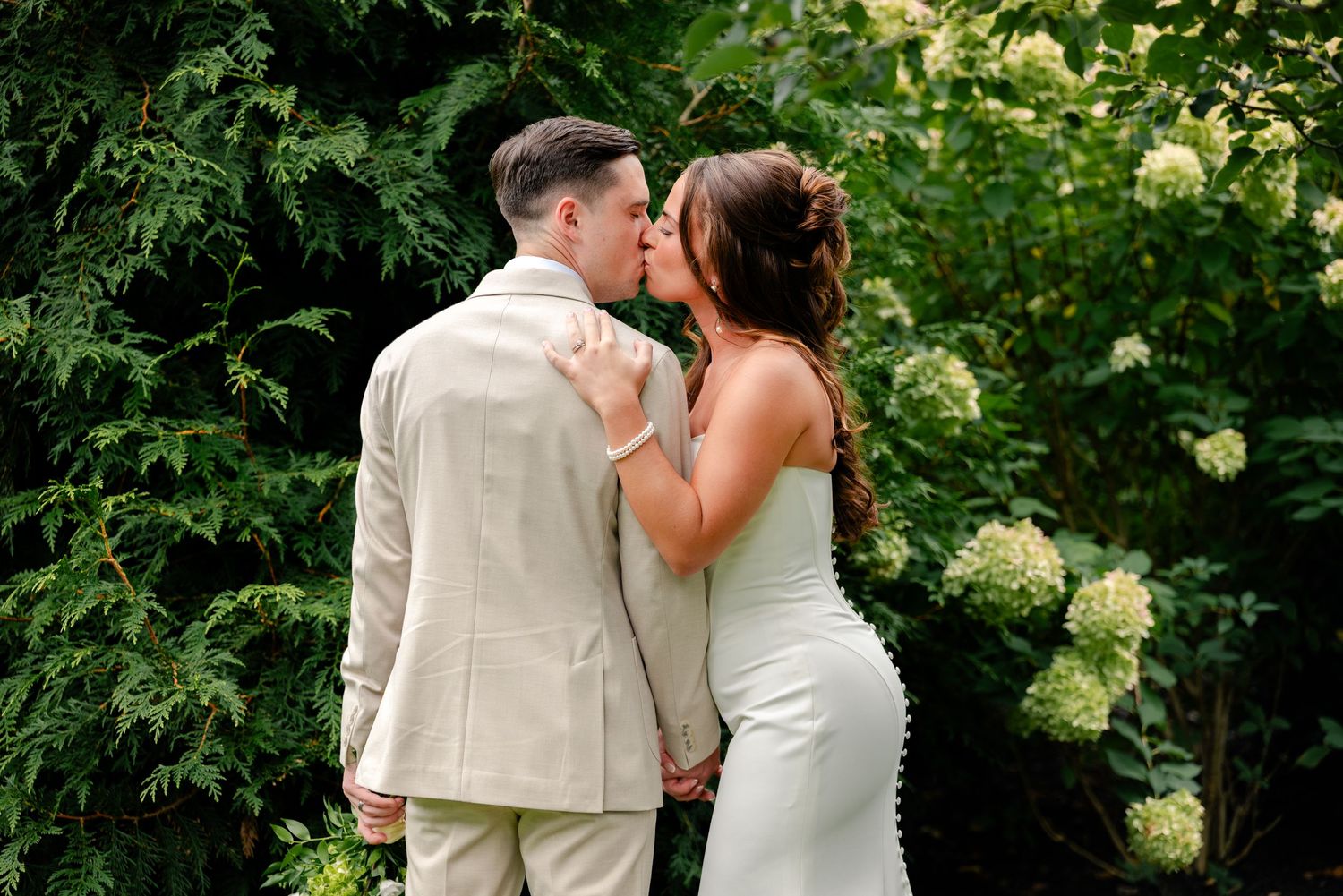 Bride in a white fitted gown and groom in a beige suit share a kiss in front of lush green foliage and white hydrangeas at their wedding.