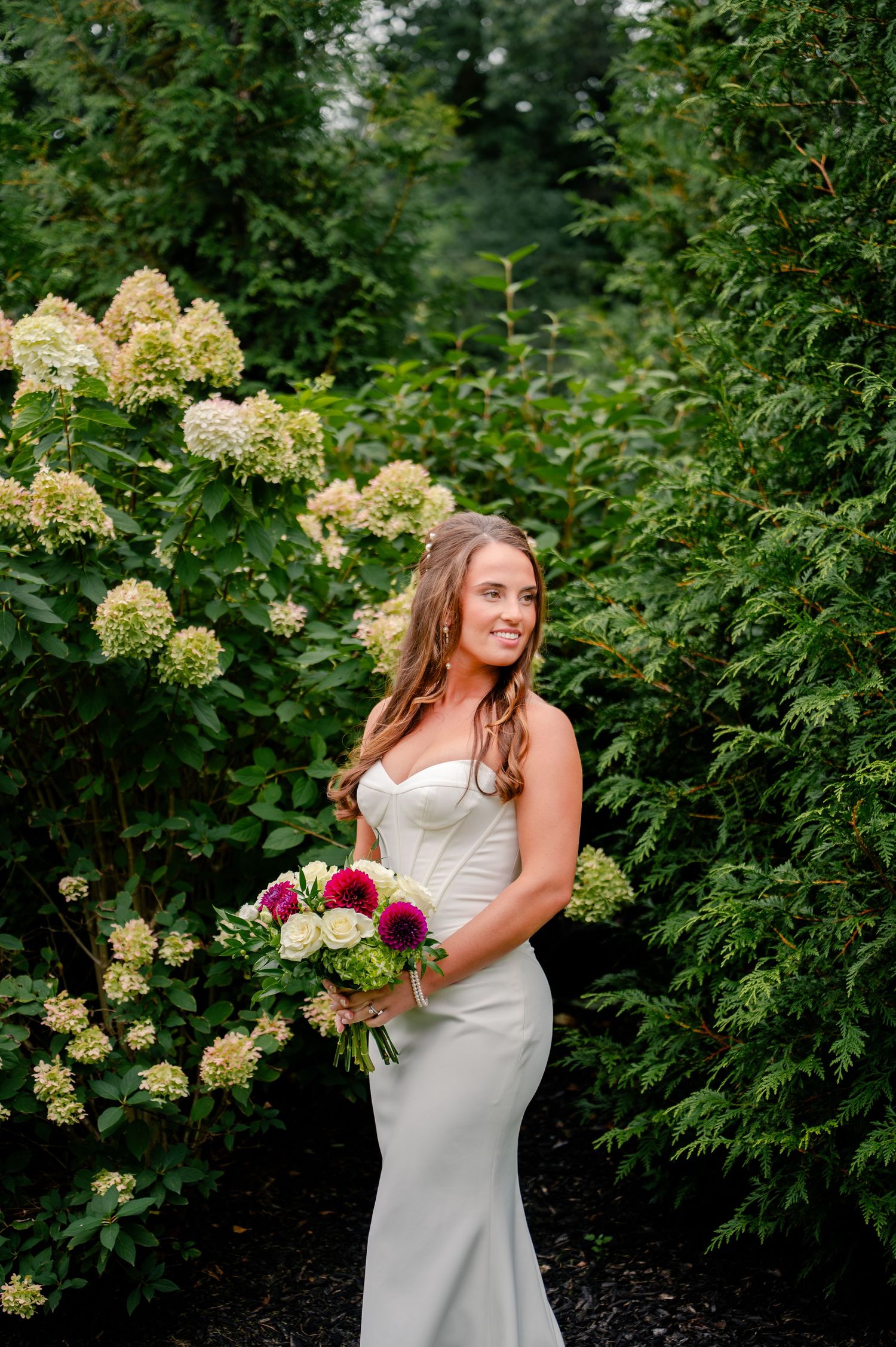 Smiling bride in a fitted white satin gown holds a pink and white bouquet beside blooming hydrangeas and evergreen hedges.