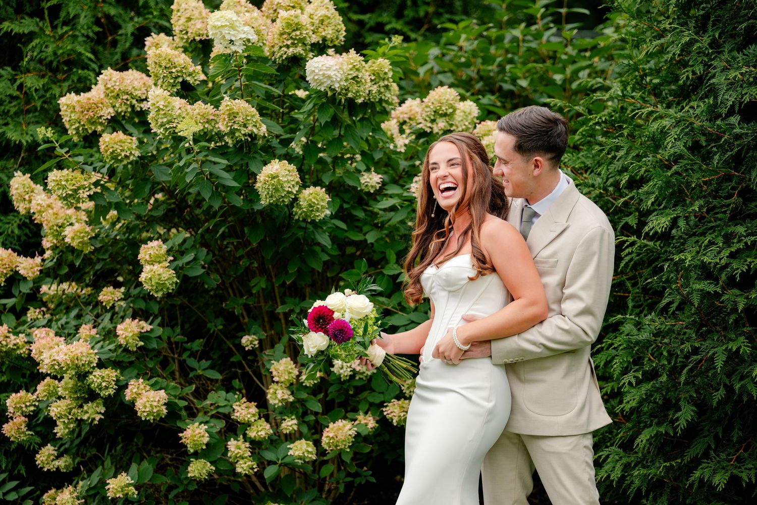 Laughing bride in a white gown holding a bouquet embraces her groom beside blooming white hydrangeas in a lush garden wedding portrait.