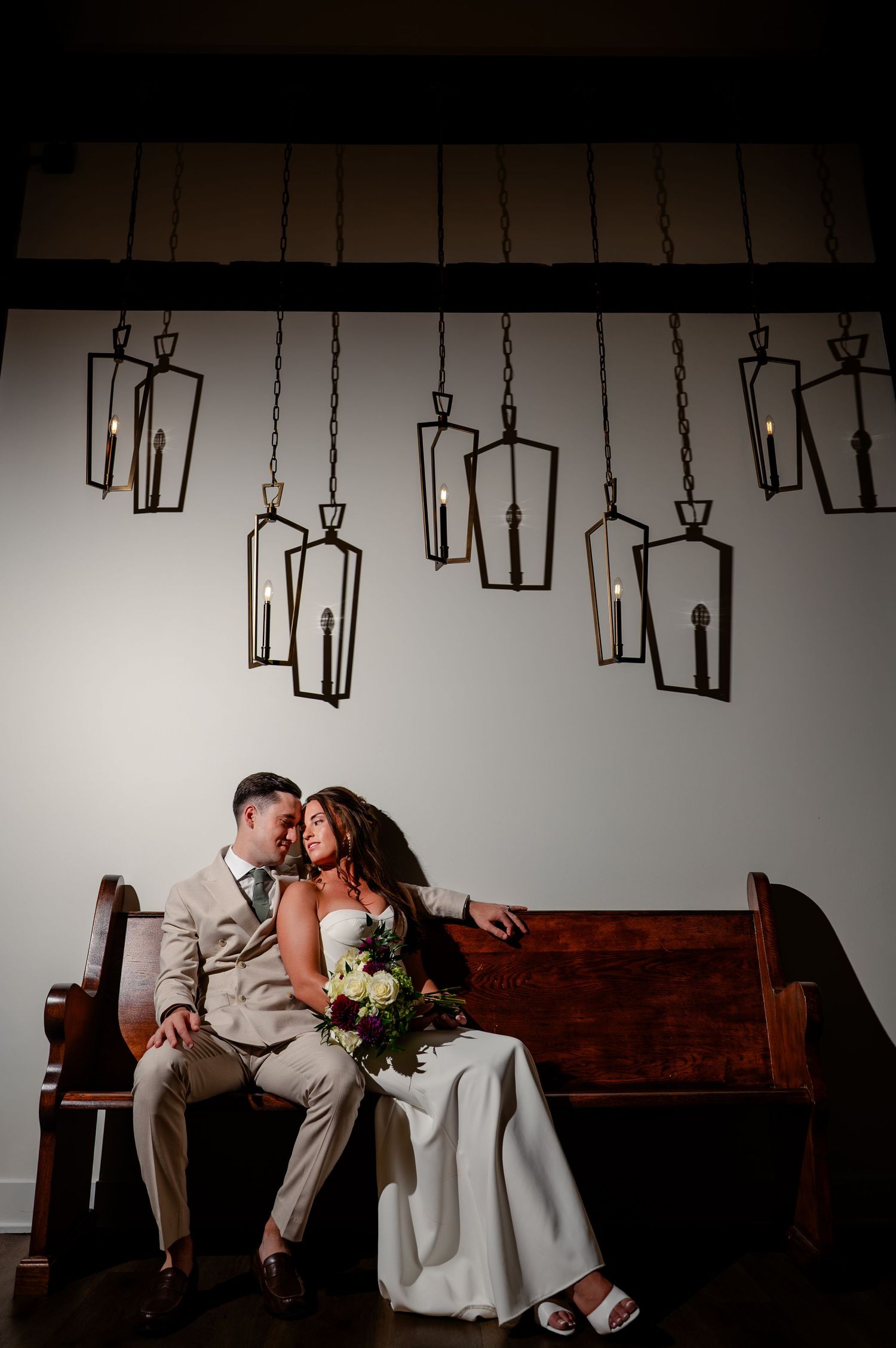 Bride and groom in a tan suit and white gown sit close on a wooden bench beneath hanging lantern lights, holding a bouquet.