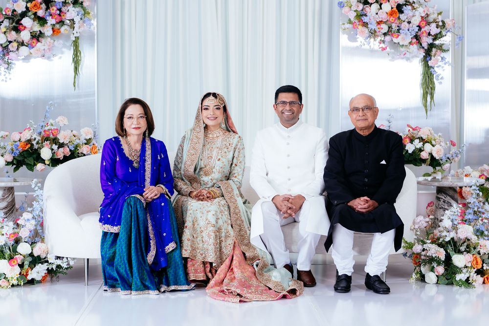 Four people in elegant South Asian wedding attire pose together on a white sofa surrounded by colorful floral arrangements.