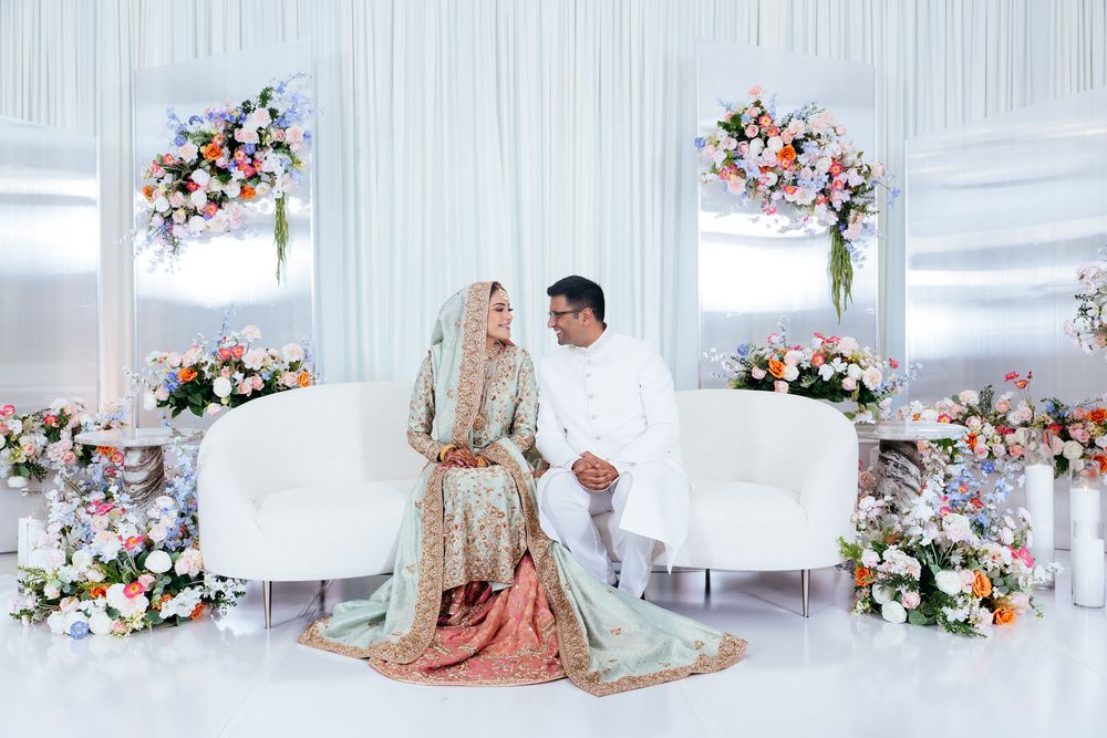 Bride and groom in traditional South Asian wedding attire sit on a white sofa surrounded by pink and white floral arrangements.