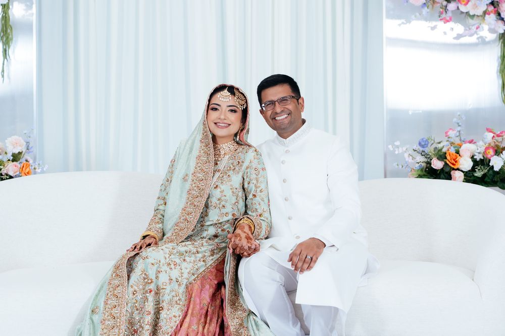 Smiling bride in an embroidered pastel green and coral outfit with a dupatta sits beside a groom in a white kurta on a white sofa.