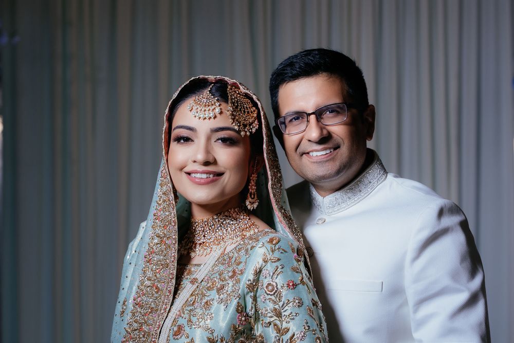 Smiling bride in an ornate teal embroidered outfit with gold jewelry poses beside a groom in a white sherwani.