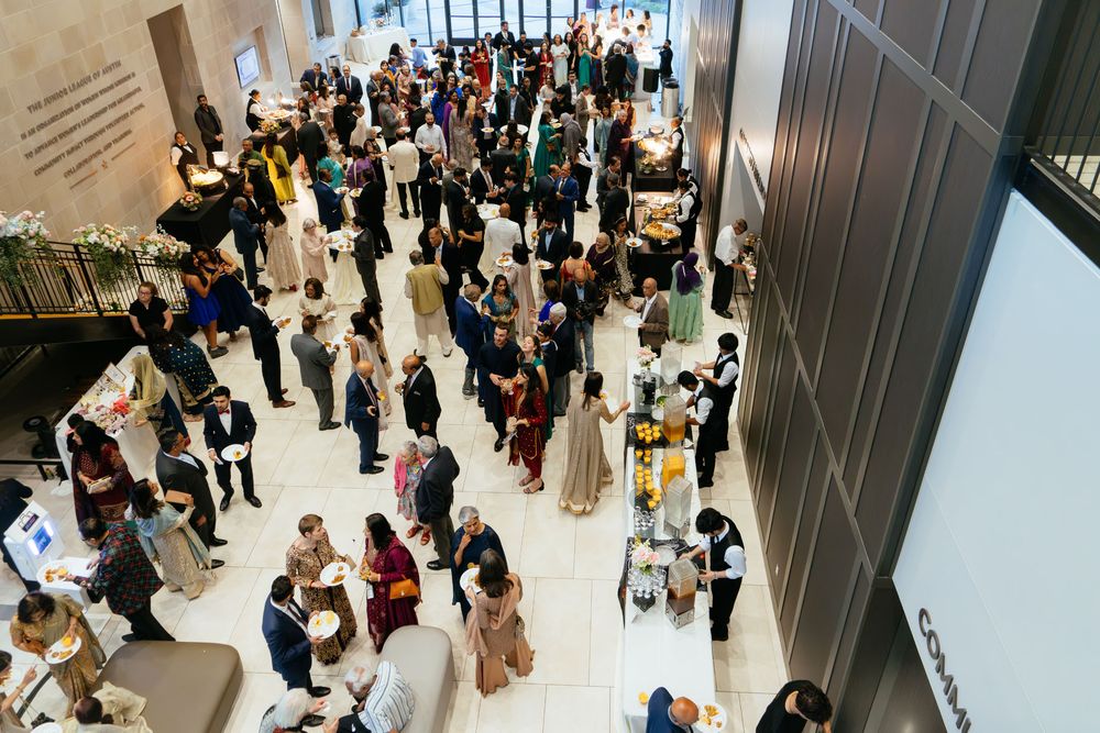 Overhead view of a large crowd of guests in formal attire mingling at a reception in a modern atrium with food and drink stations.