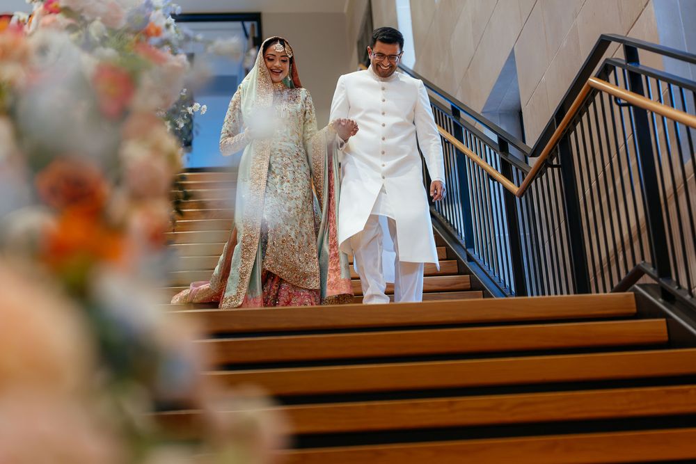 Bride in an ornate embroidered gown and veil walks down wooden stairs beside a groom in a white kurta, with flowers in the foreground.