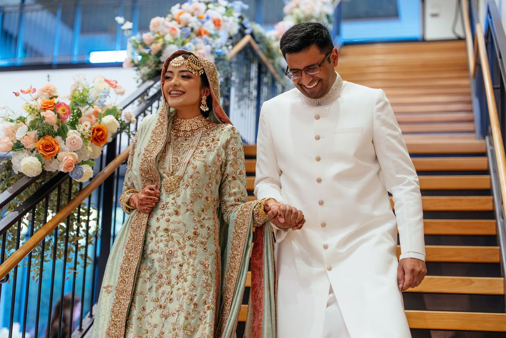 A smiling couple in elegant South Asian wedding attire holds hands on a staircase beside lush floral arrangements in peach, white, and pink.