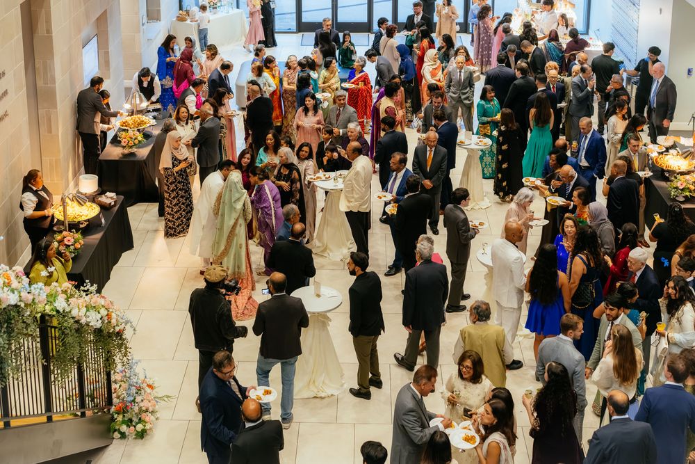 Guests in formal and traditional attire mingle at an elegant reception in a bright atrium with floral arrangements and buffet tables.
