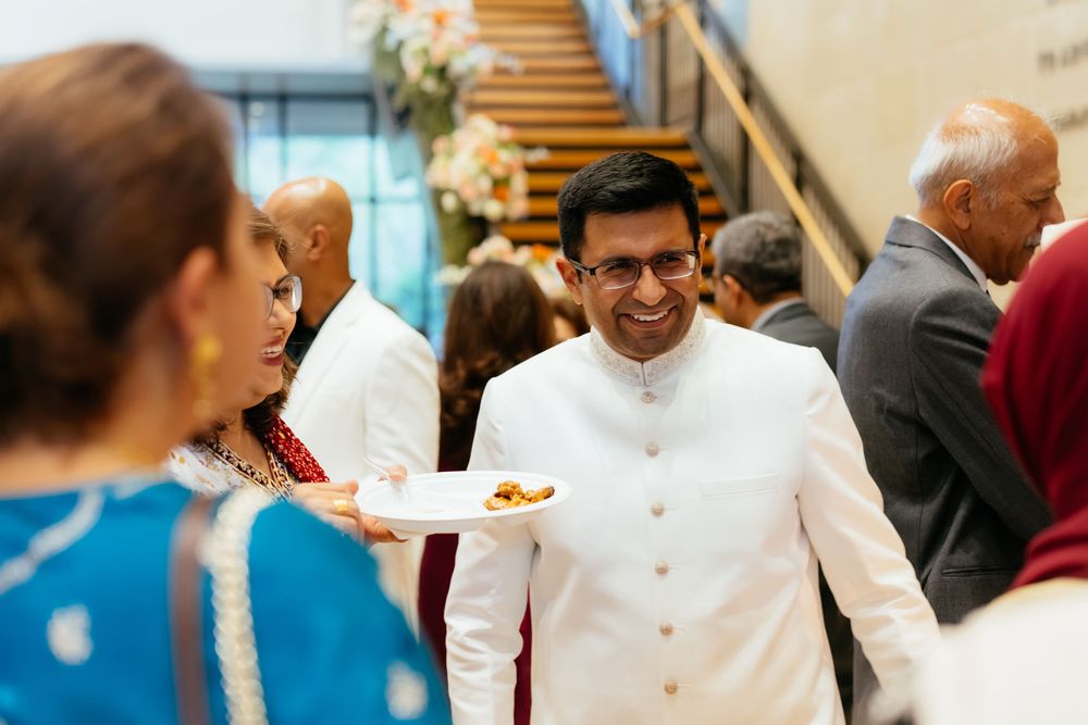 Smiling man in a white sherwani holds a plate of food while mingling with guests at a festive wedding gathering.