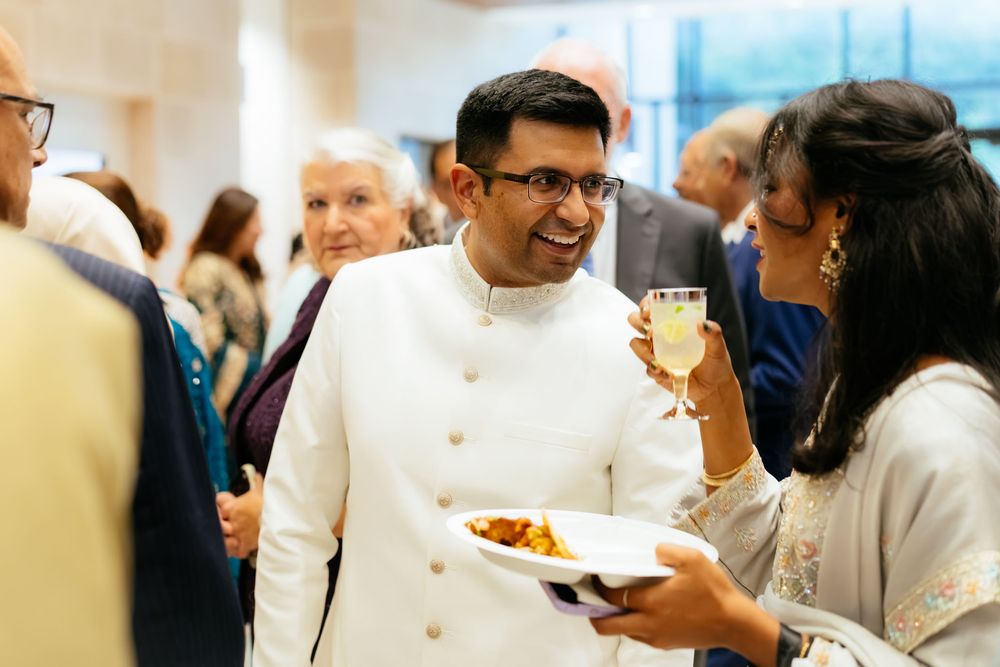 Smiling man in a white ivory jacket holds a plate of appetizers while guests chat and mingle at an elegant indoor reception.