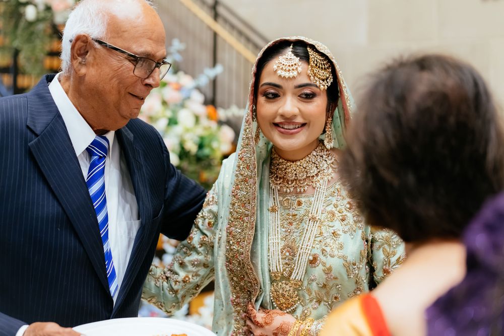 Smiling bride in an ornate green and gold embroidered South Asian wedding outfit and jewelry stands beside an elderly man in a navy suit.
