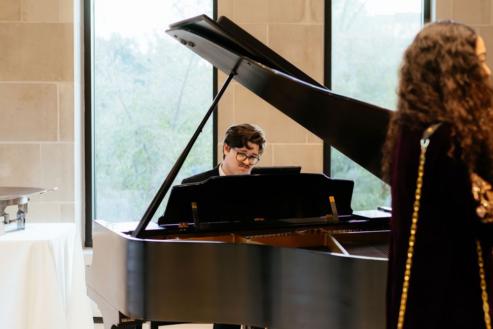 A pianist in glasses plays a black grand piano in a bright stone-walled hall while a woman with curly hair watches.