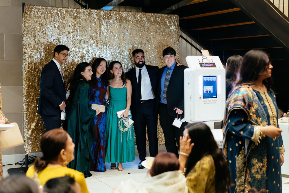 Guests in formal and traditional attire pose for a group photo near a photo booth in front of a gold sequin backdrop.