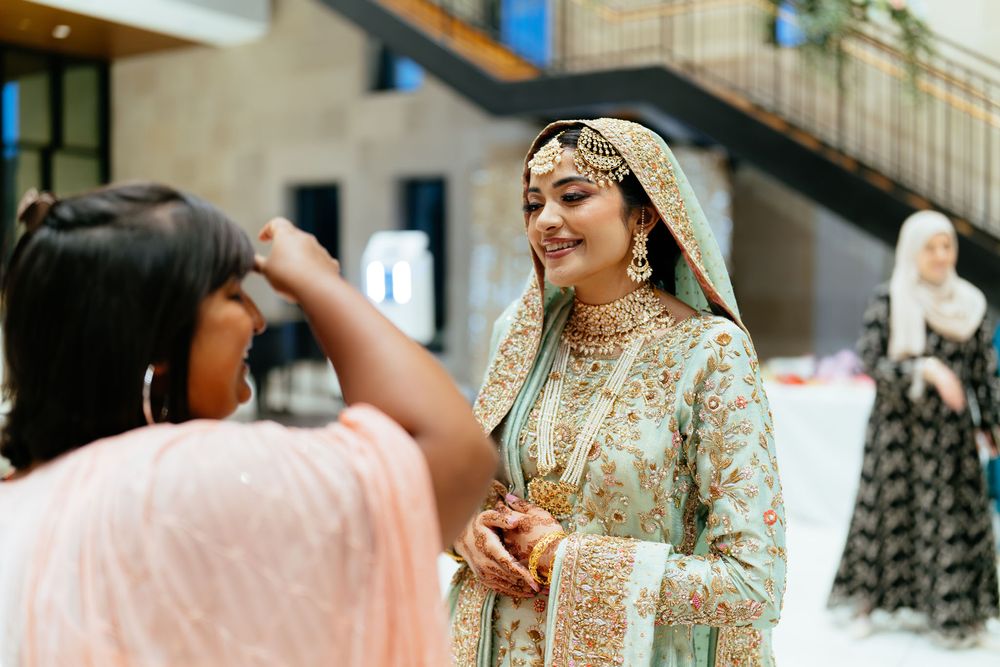 Smiling woman in an ornate mint green embroidered South Asian outfit with gold jewelry adjusts her look while a friend in pink watches.