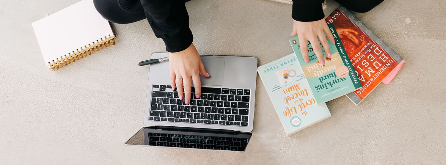 Overhead view of a person sitting on the floor typing on a silver laptop beside a spiral notebook and a stack of colorful books.