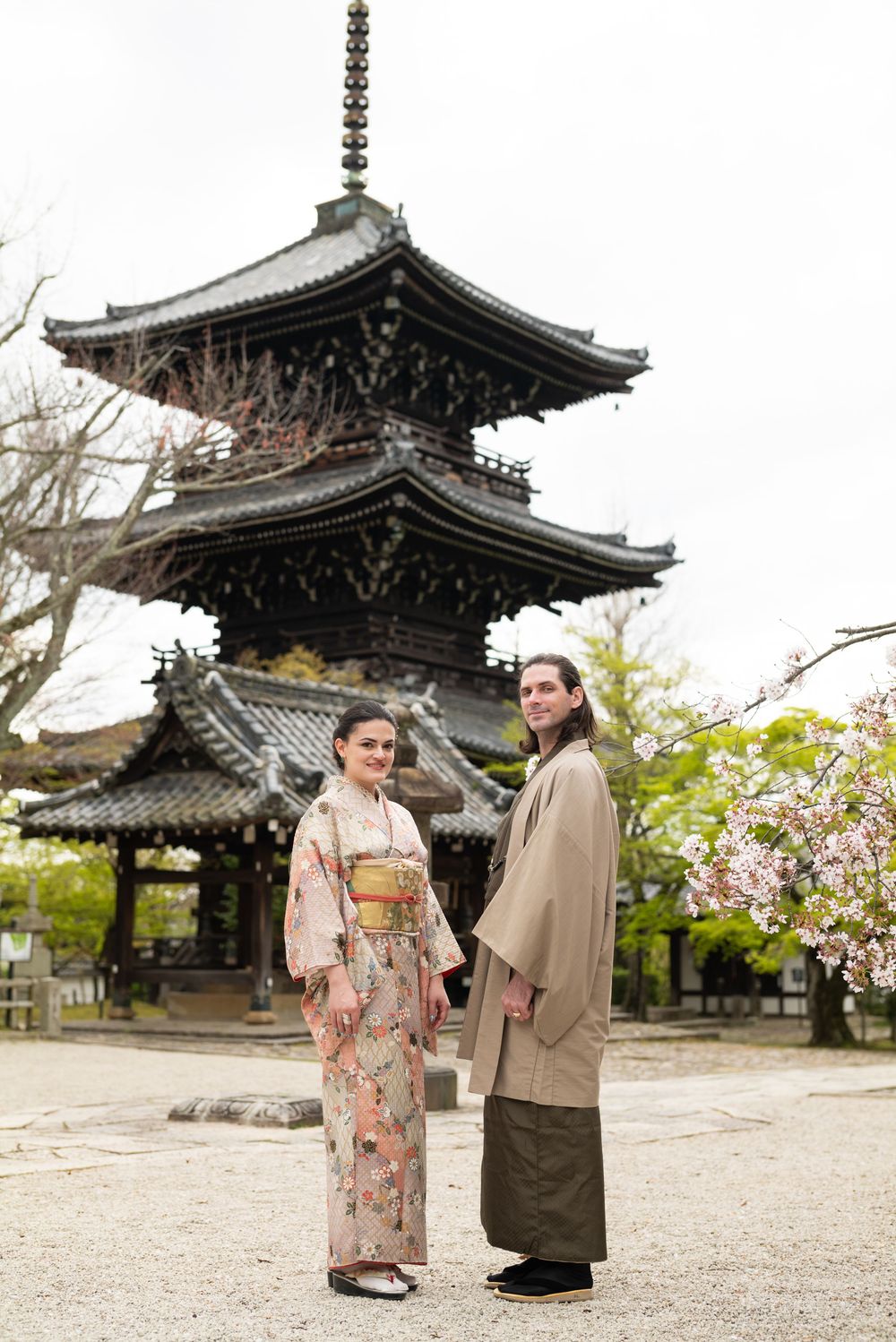 Couple in traditional Japanese kimono and haori standing hand in hand before a historic wooden pagoda beside blooming cherry blossoms.