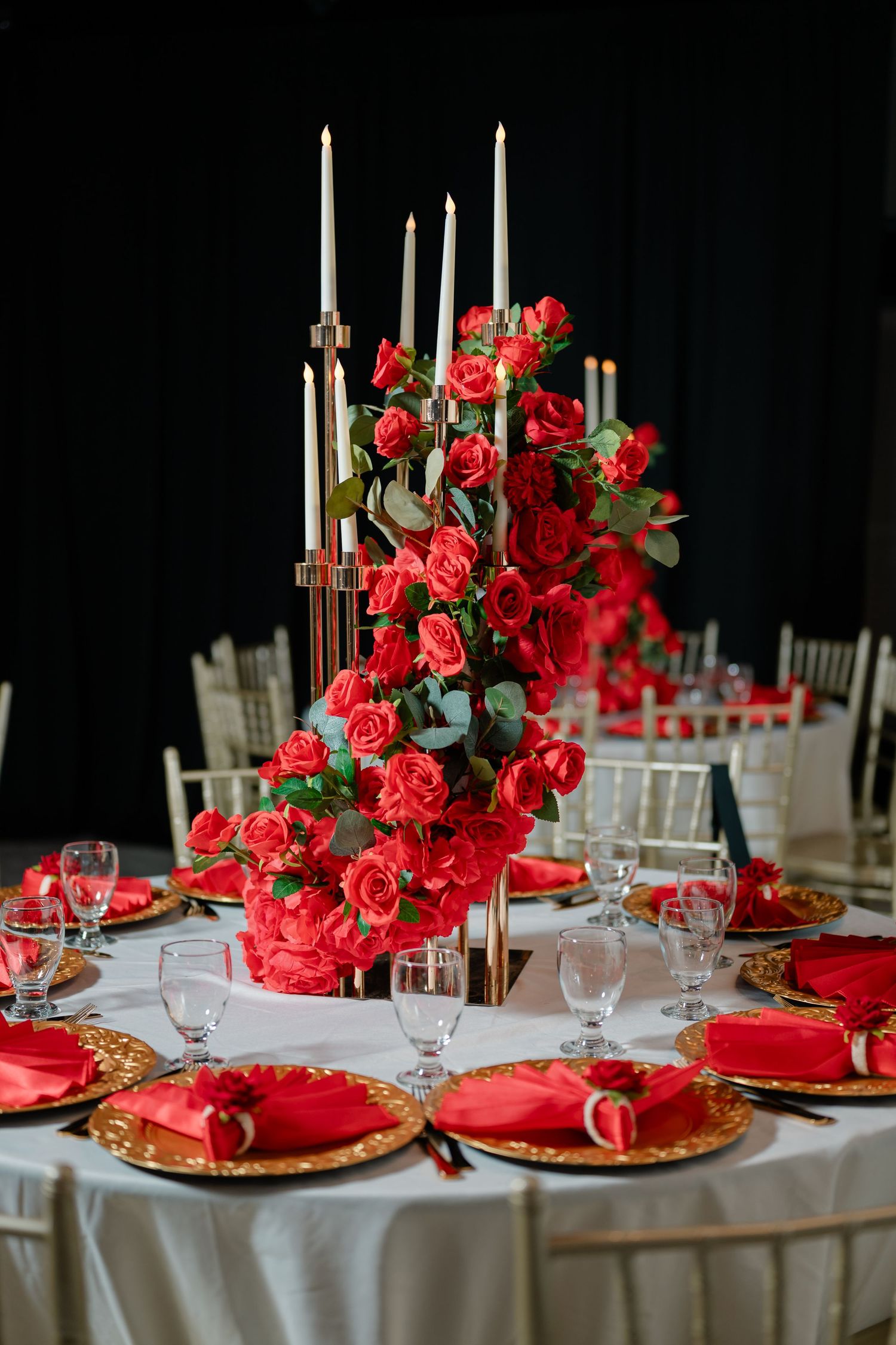 Elegant wedding reception table with a tall red rose centerpiece, white taper candles, gold charger plates, and red napkins.