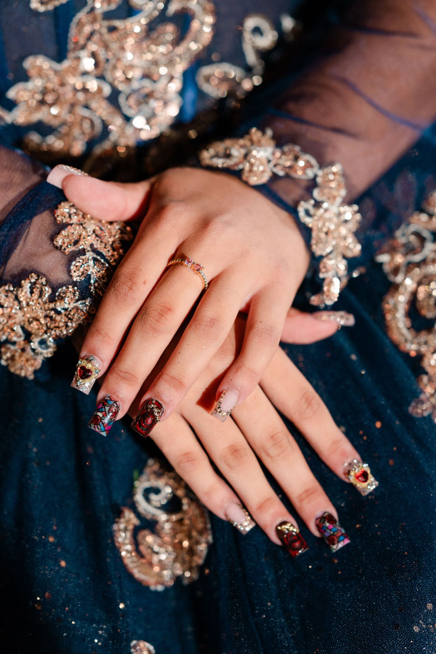 Hands with ornate red and glitter nail art and a gold ring rest on a navy blue tulle gown embellished with gold sequined embroidery.