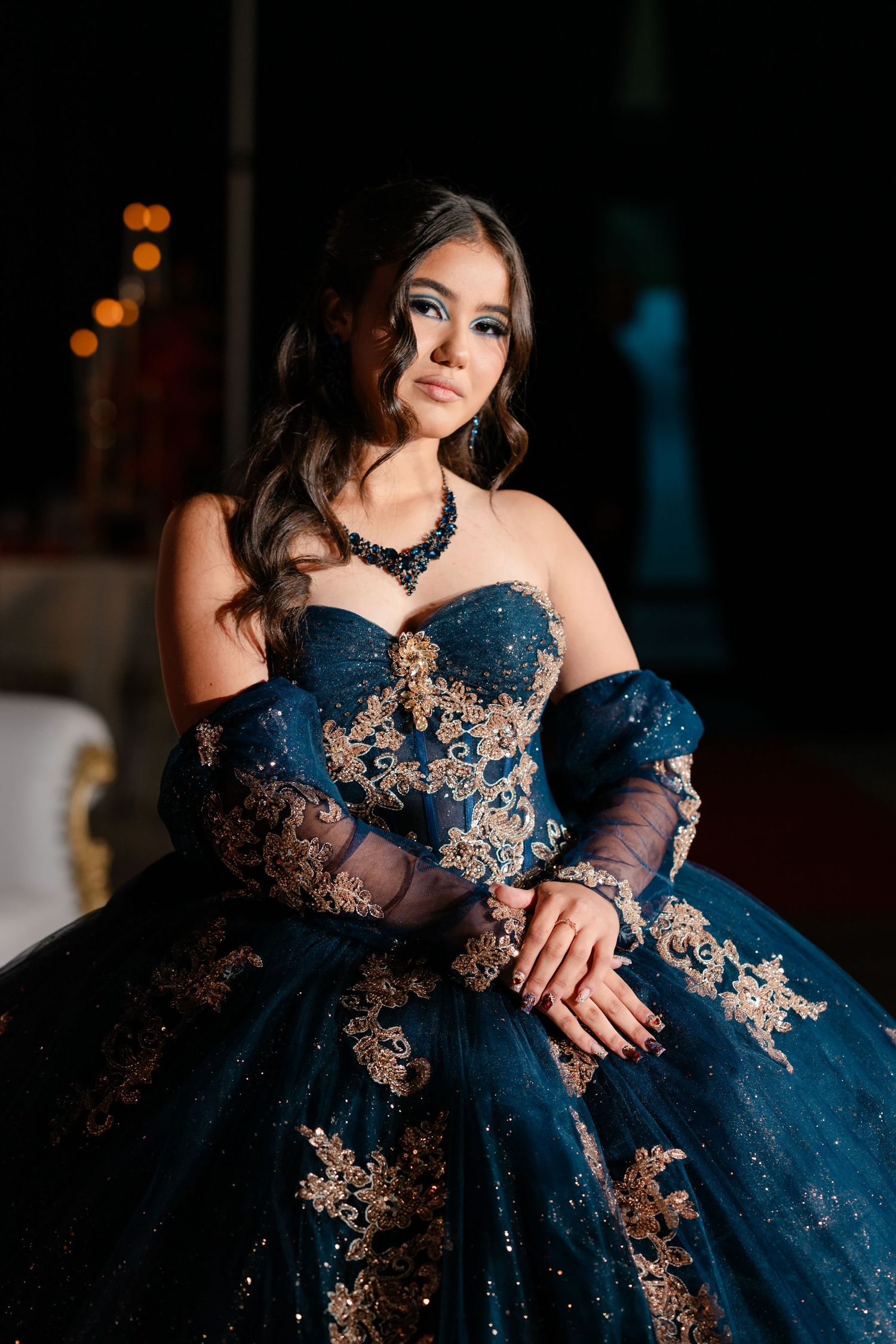 Young woman in a navy blue quinceañera ball gown with gold lace appliqués, sheer sleeves and a jeweled necklace posing elegantly.