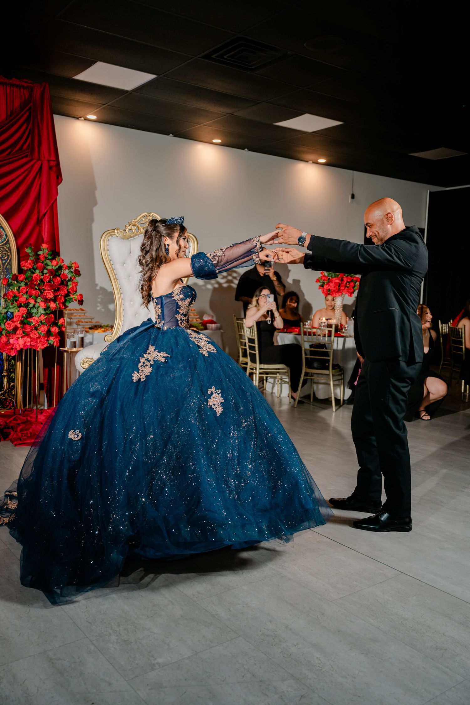 Quinceañera in a navy blue ball gown with gold appliqués dances with a man in a black suit beside a gold throne and red floral decor.