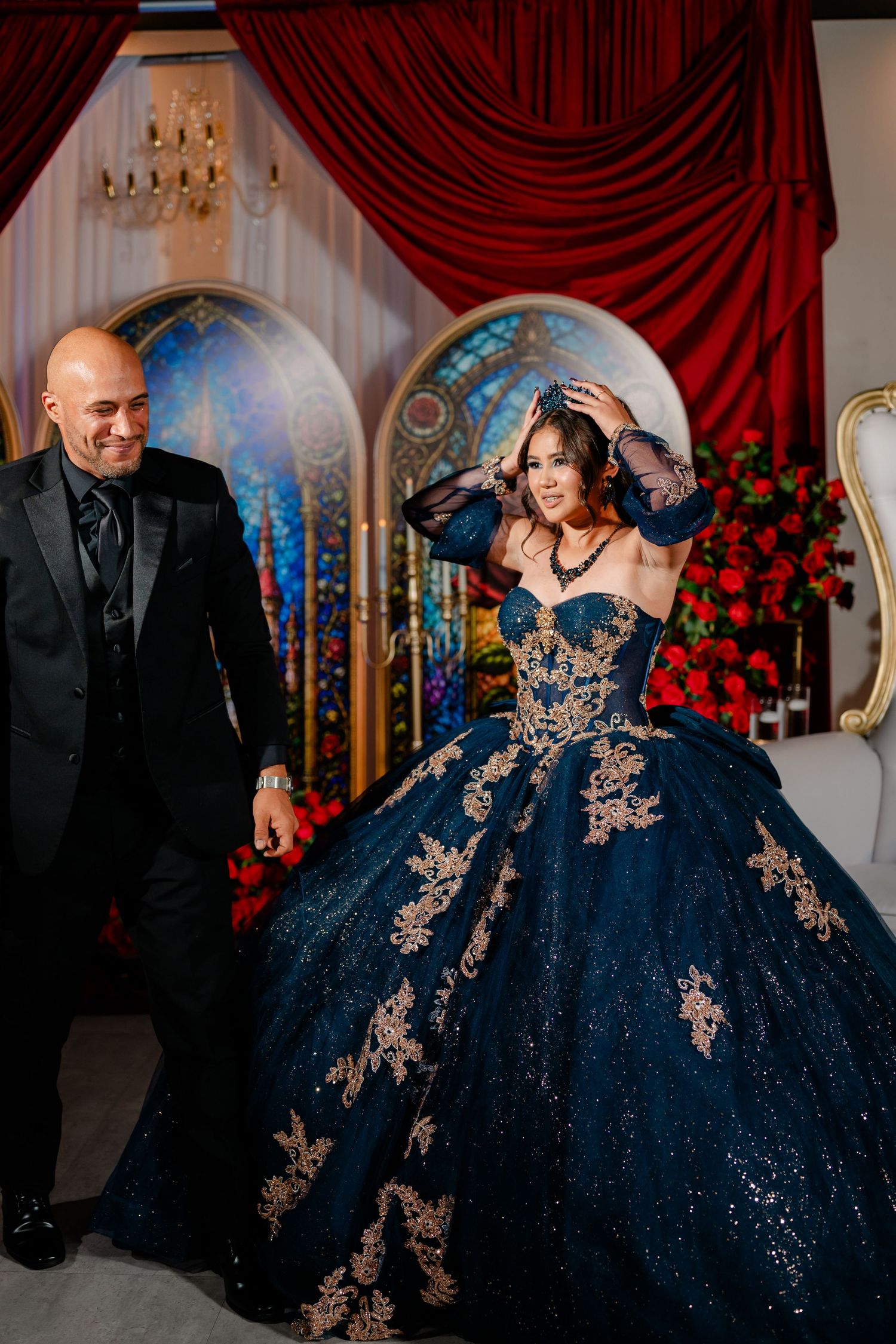 Smiling man in a black suit stands beside a woman in a navy blue quinceañera ball gown with gold appliqué, framed by red drapes and staine