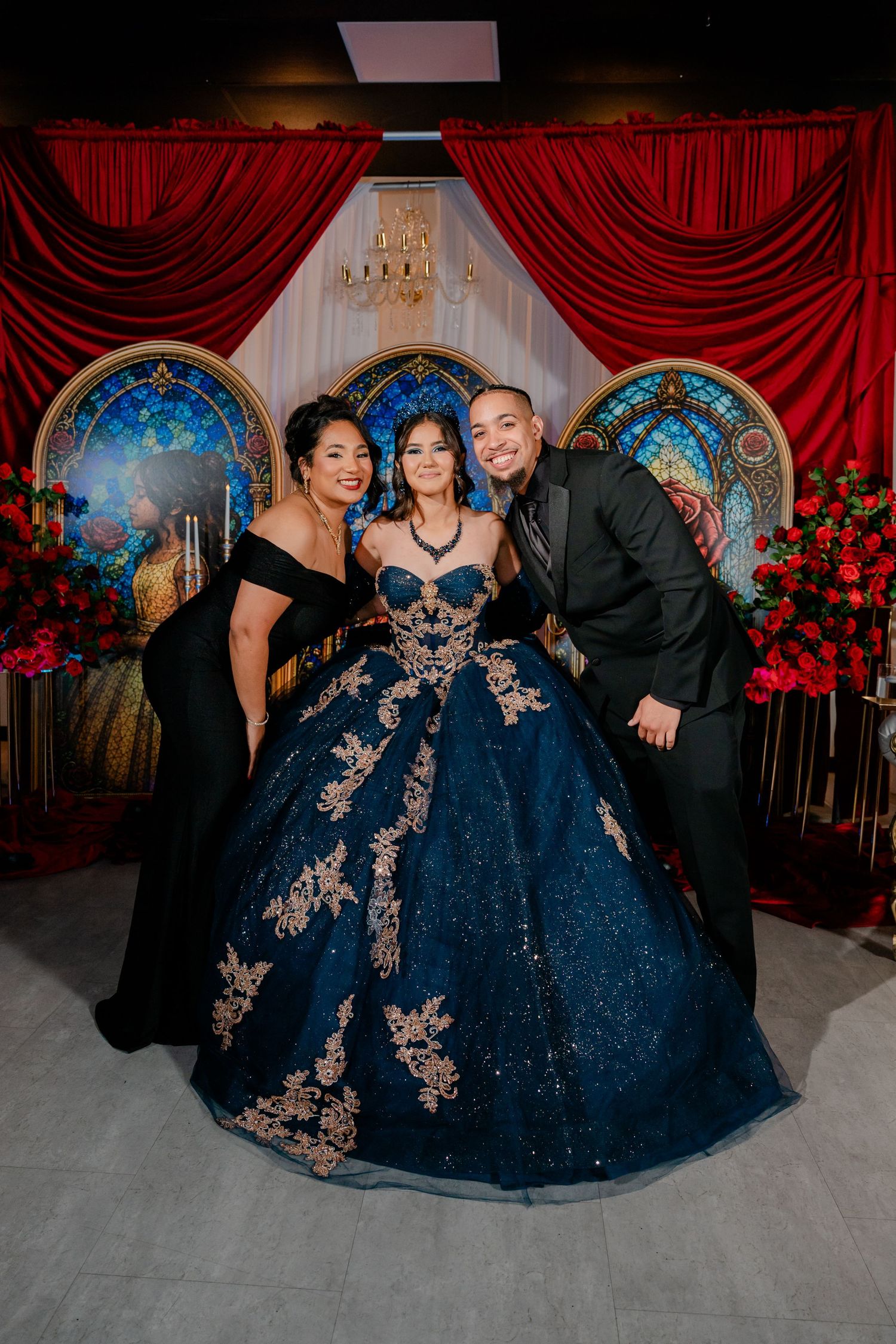 Quinceañera in a navy blue gold-embroidered ball gown poses with a woman and man in black before red drapes and stained glass.