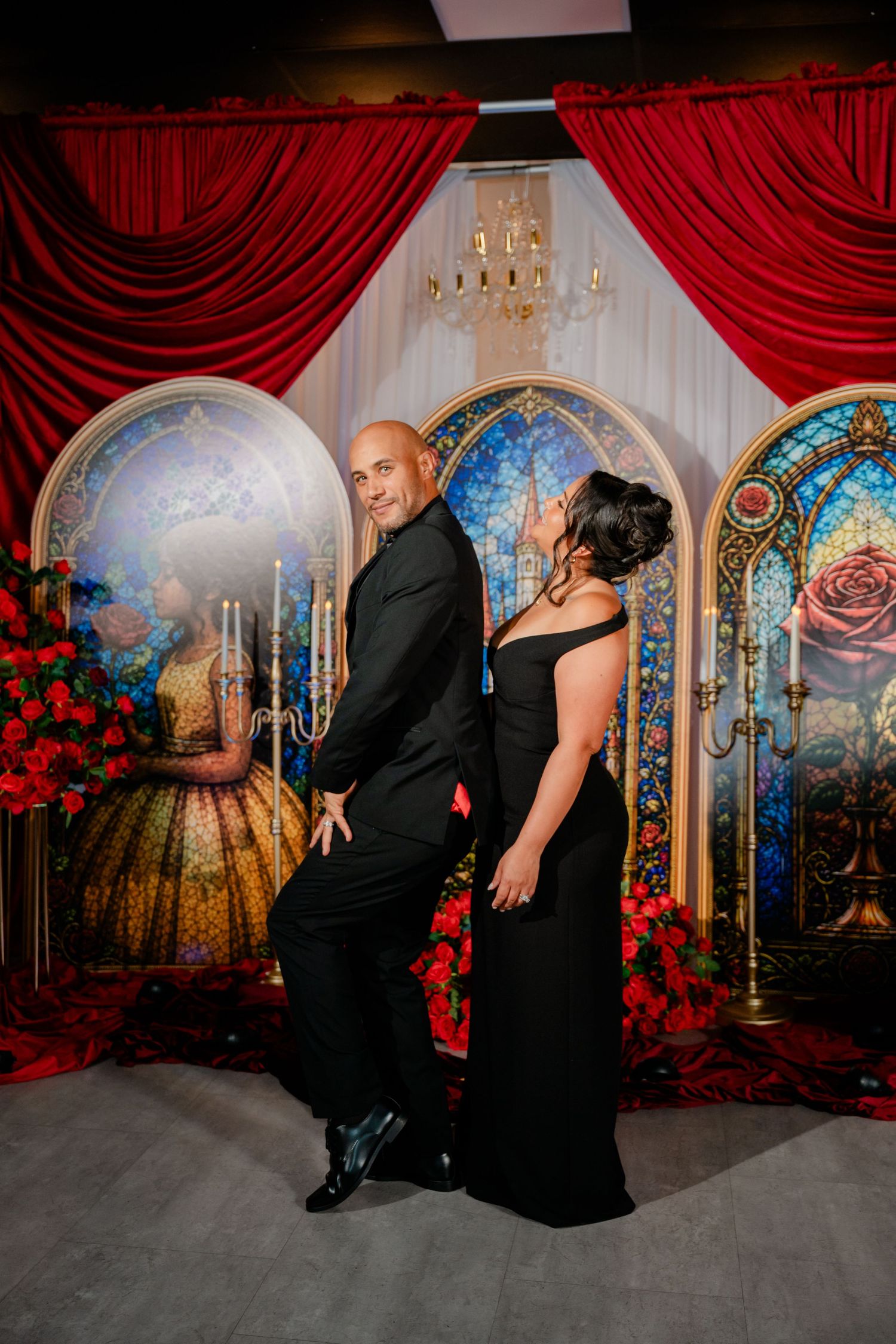 Couple in black formal attire pose in front of red curtains, stained glass arches, and roses at a fairytale-themed event.