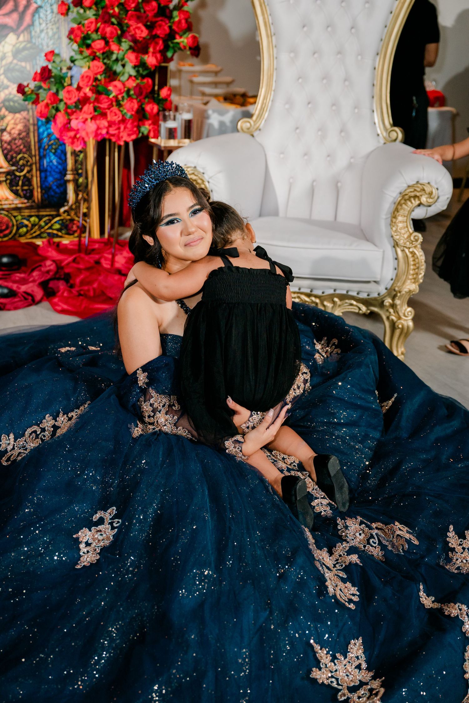 Woman in an embellished navy quinceañera gown and tiara hugging a small child in black beside an ornate white throne chair and red roses.