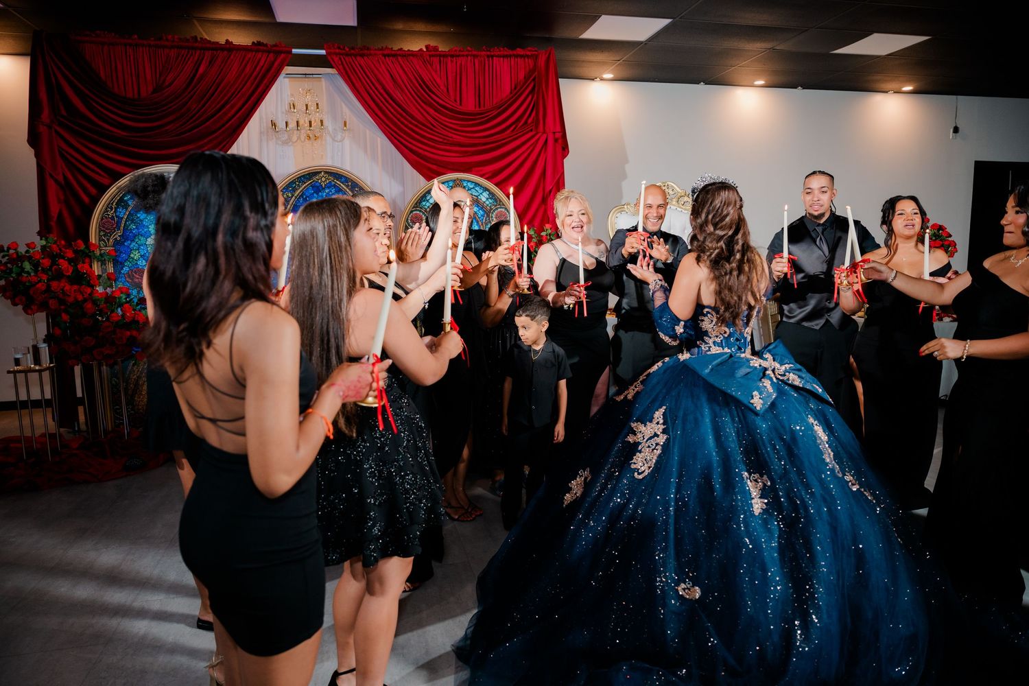 Guests gather around a girl in a sparkling blue quinceañera ball gown at an elegant celebration with red draped curtains.