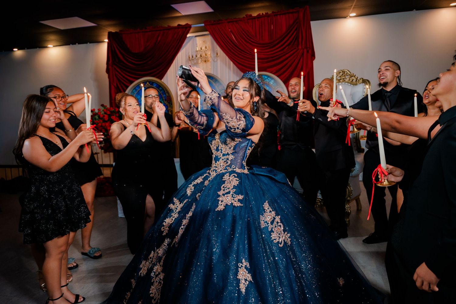 Guests holding candles celebrate around a young woman in a sparkling navy blue quinceañera ball gown at a festive party with red drapes.