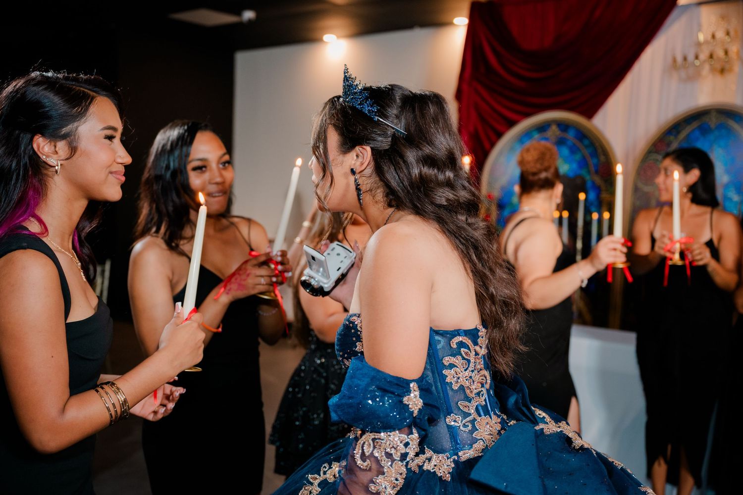 Quinceañera in a blue embroidered gown and tiara holds a candle while smiling guests in black dresses gather around during the ceremony.