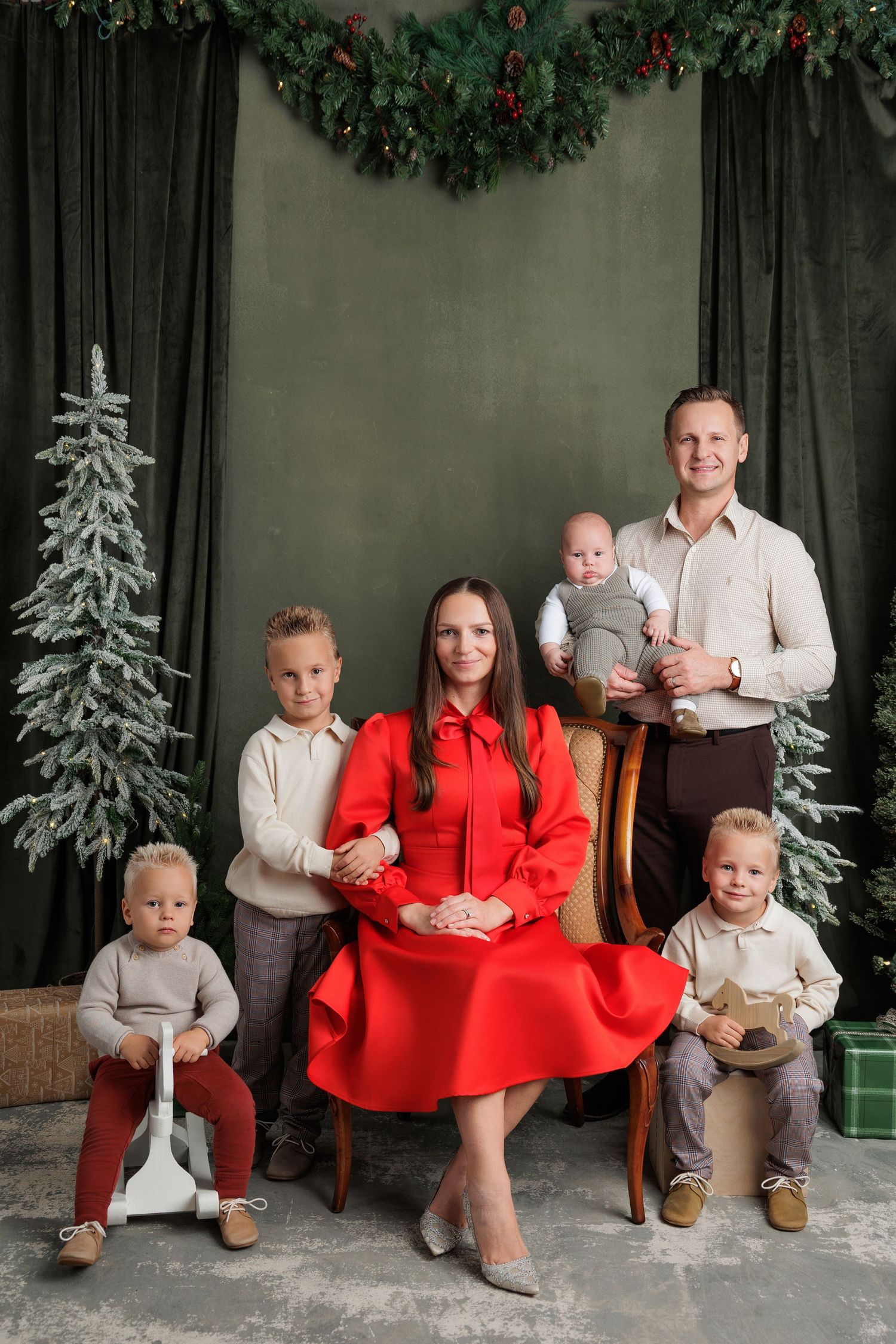 Family Christmas portrait of parents and four young children posed by a snowy tree and green garland, mother in a red dress seated.