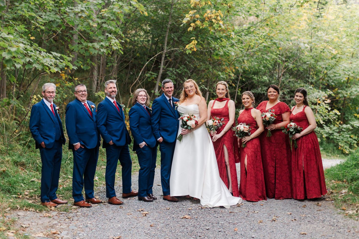 Bride in white gown stands with groomsmen in navy suits and bridesmaids in red dresses holding bouquets on a gravel path in the woods.