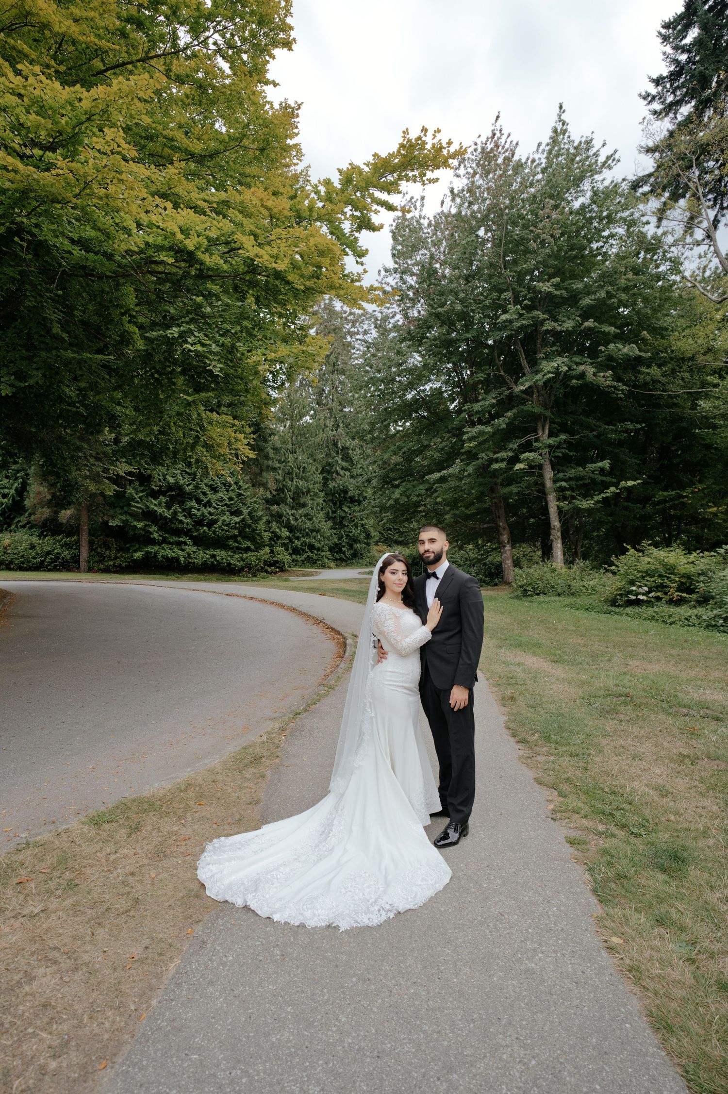 Bride in a long lace wedding gown and groom in a black suit pose together on a curved park path surrounded by tall green trees.