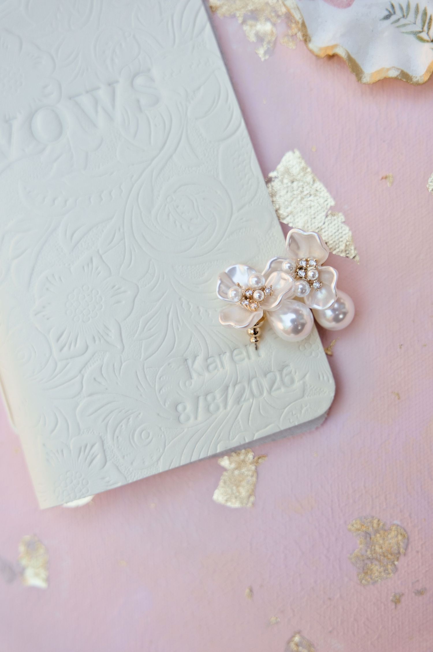 Cream embossed floral wedding vows booklet dated 8/8/2026 with pearl and blossom earrings on a pink background with gold leaf accents.