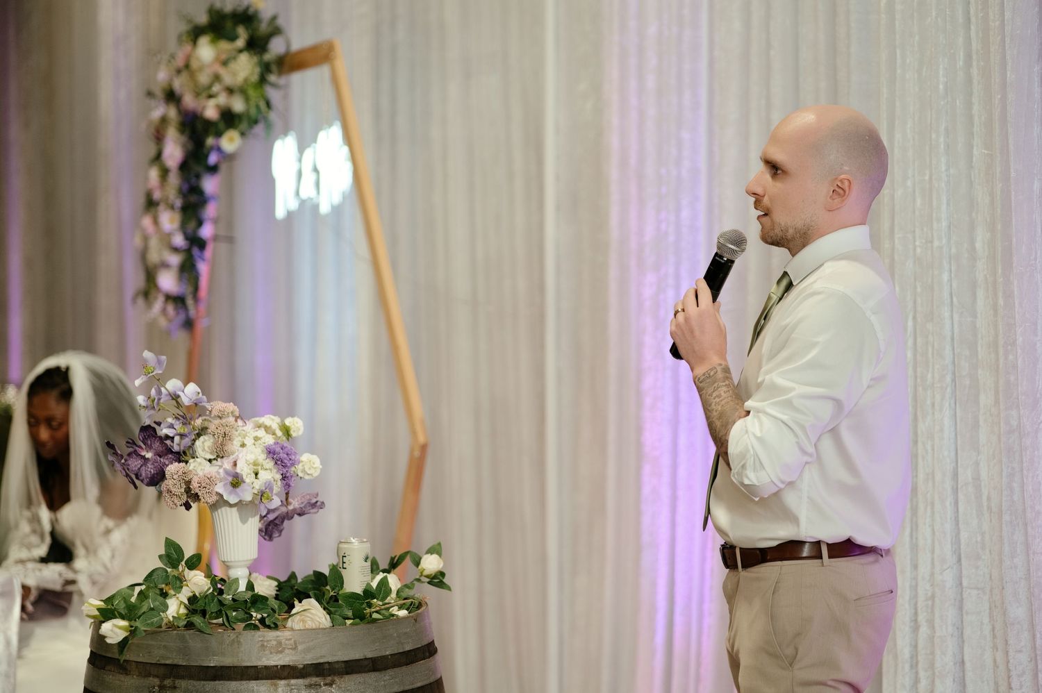 Groom in a white shirt and khaki pants gives a wedding speech into a microphone beside a floral arrangement while the veiled bride looks on.