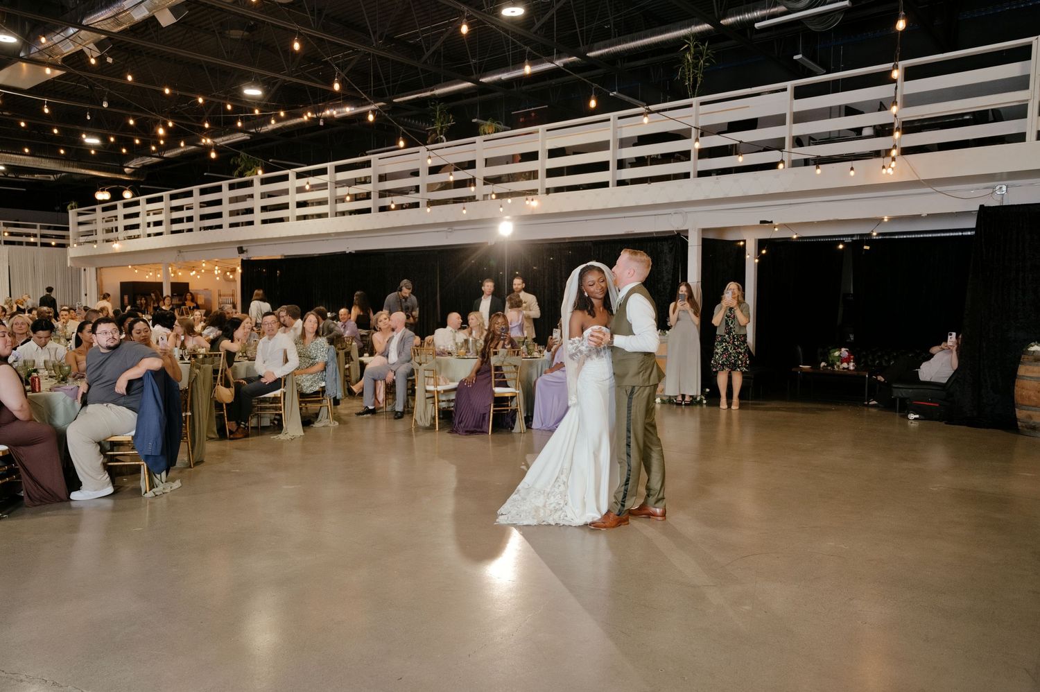 Bride and groom share a first dance on a polished floor while seated wedding guests watch inside a rustic venue with string lights.