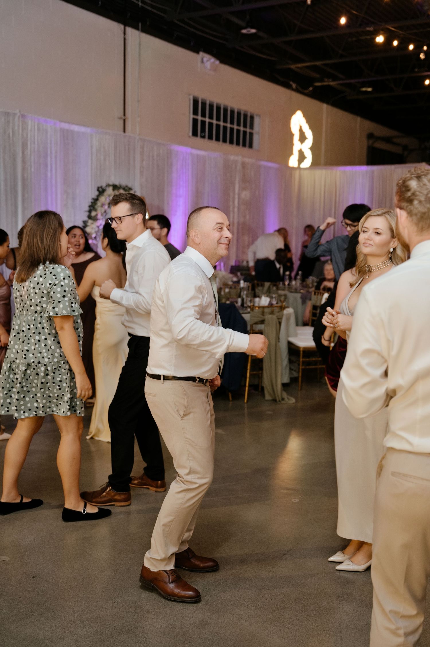 Wedding guests in dressy attire dance on a concrete floor at a reception with purple uplighting and sheer draped curtains.