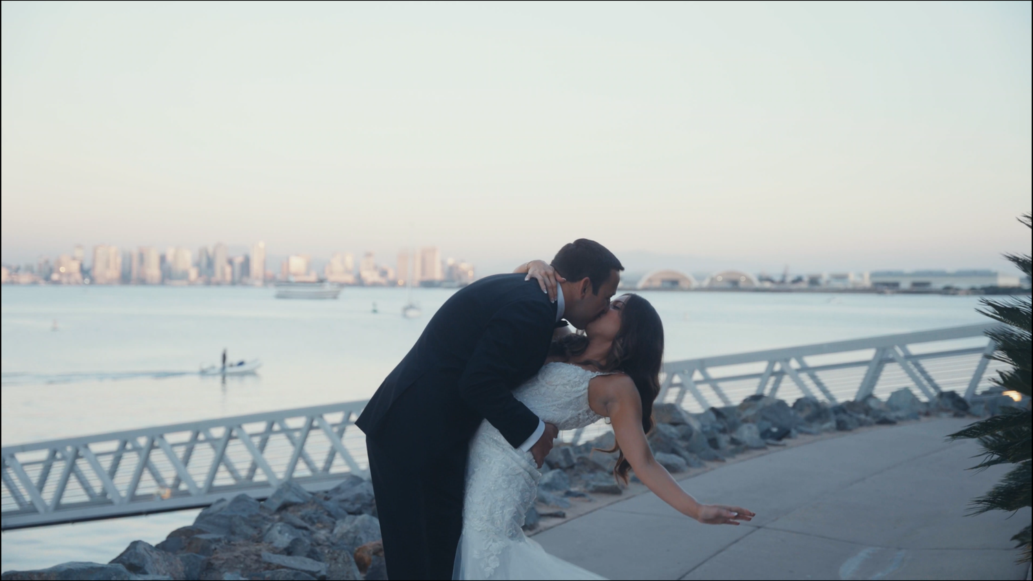 A bride and groom share a romantic dip kiss by the waterfront with a city skyline at sunset.