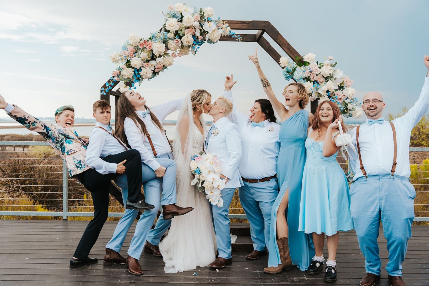 Wedding party celebrating under floral arch, bridesmaids in blue dresses, groomsmen in suspenders, bride and groom kissing.
